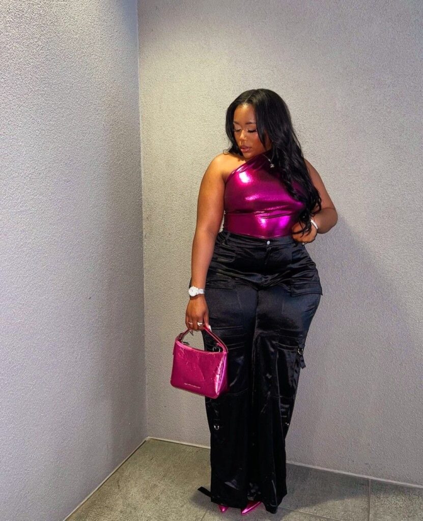 Black plus size woman in a shiny pink top and satin black cargo pants with pink heel and a hot pink purse.