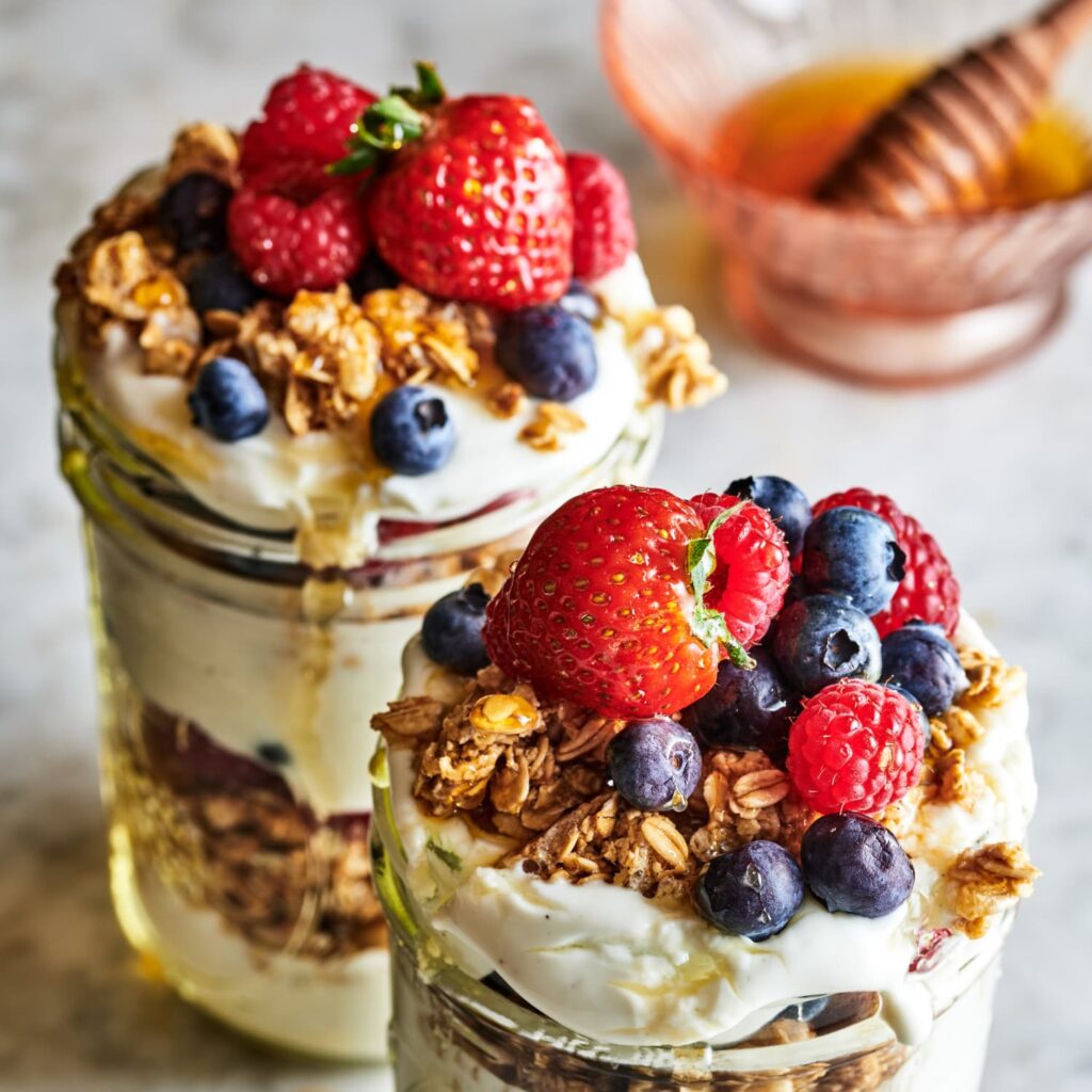 Two fruit parfait cups, topped with strawberries, blueberries, and rasberries, with granola and honey.