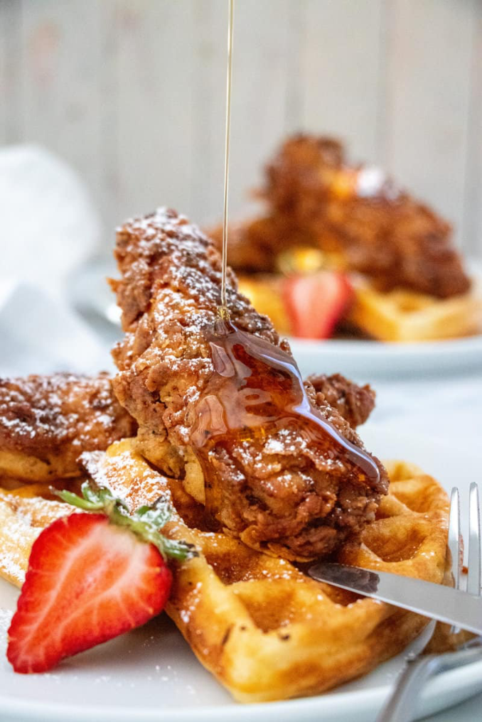 A fried chicken cutlet laid across a Belgian waffle, drizzled with maple syrup with a side of strawberries.