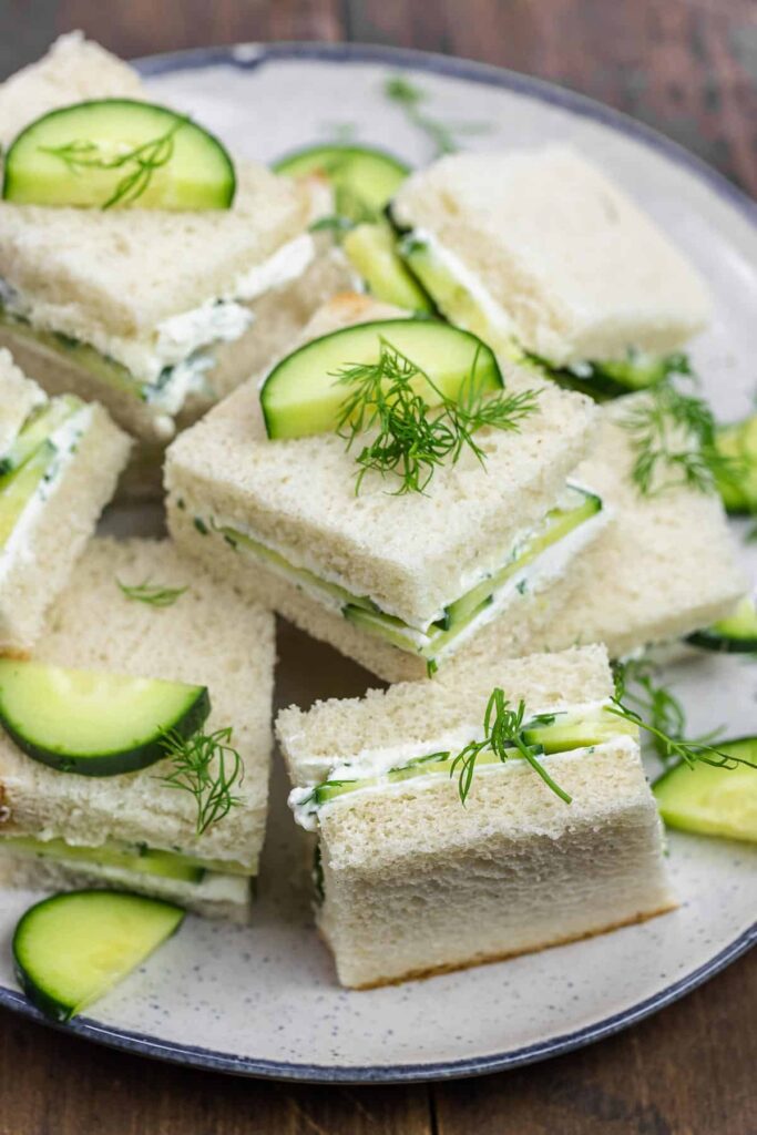 A bunch of cucumber sandwiches stacked on top and around each other with added cucumber slices and sprigs of fresh herbs